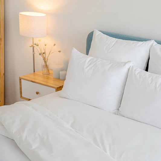 Photo of white fitted cooling sheet and cooling pillow cases made up on bed with nightstand, lamp and floral arrangement